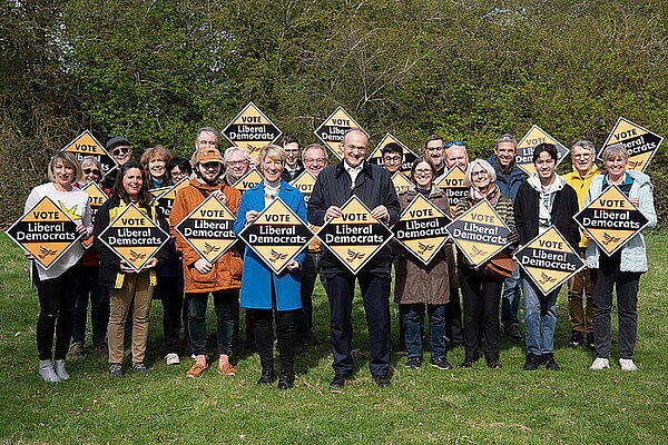 A crowd of Liberal Democrats hold Liberal Democrat gold diamond shaped posters behind their newly selected parliamentary candidate, Pippa Heylings