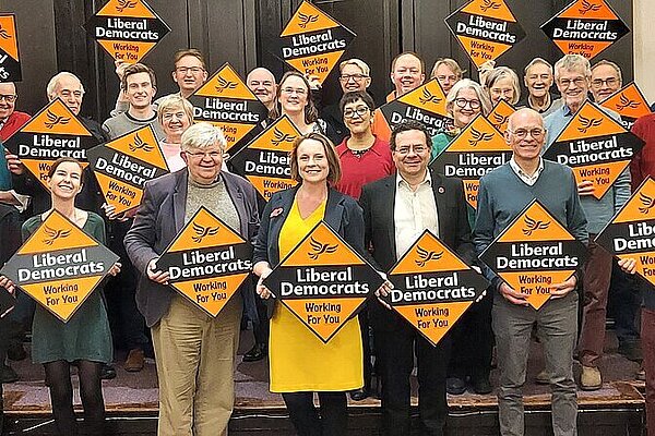A crowd of Liberal Democrats hold Liberal Democrat gold diamond shaped posters behind their newly selected parliamentary candidate, Cheney Payne