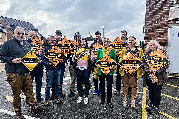 A crowd of Liberal Democrats hold Liberal Democrat gold diamond shaped posters in a car park