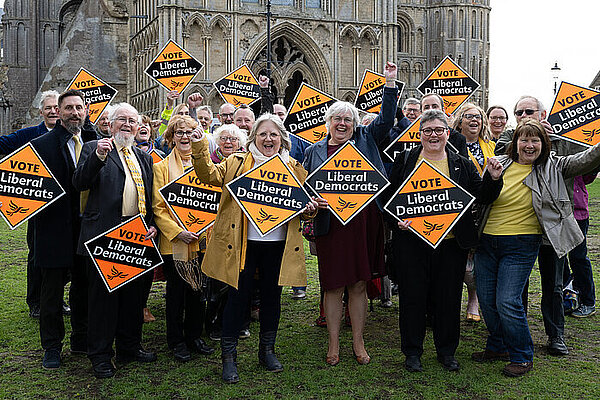 A crowd of Liberal Democrats hold Liberal Democrat gold diamond shaped posters on a grass space in front of Ely Cathedral