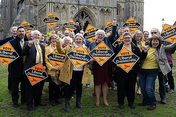 East Cambridgeshire Liberal Democrats A crowd of Liberal Democrats hold Liberal Democrat gold diamond shaped posters on a grass space in front of Ely Cathedral