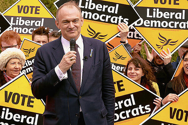 Ed Davey MP in a suit in front of a crowd of cheerful people holding large Liberal Democrat diamond posters
