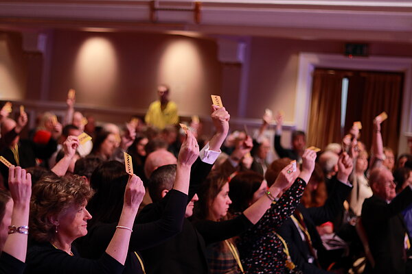 A crowd of people in a conference room many voting with their hands in the air