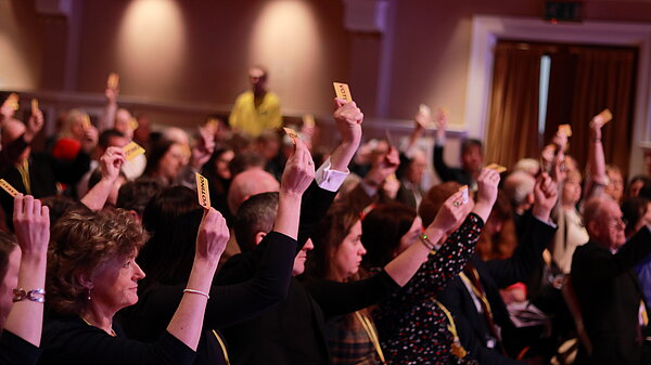 Crowd of people sat in a room many voting by raising their arms